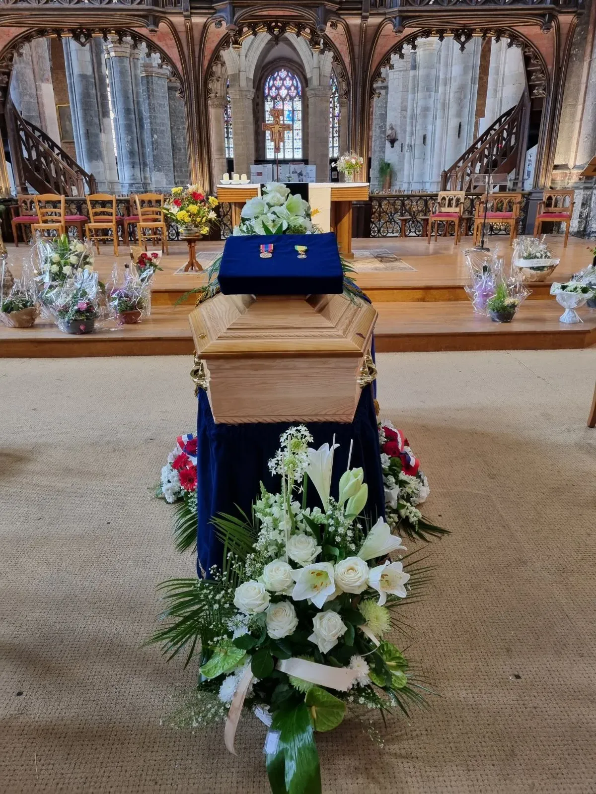 Cercueil en bois dans une église entouré de compositions florales funèbres.